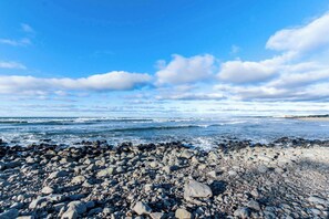 Una spiaggia rocciosa con onde, cielo sereno e edifici in lontananza.