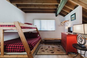 Ein Etagenbett mit rot-schwarzem Karomuster, ein Schreibtisch mit Lampe, ein Fernseher und ein Fenster mit Jalousien.