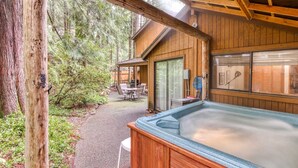 Eine Holzhütte mit Whirlpool, einer Terrasse und Gartenmöbeln, vor einer bewaldeten Kulisse.