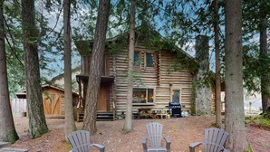 Une cabane en rondins avec trois fauteuils Adirondack devant, entourée de grands arbres.