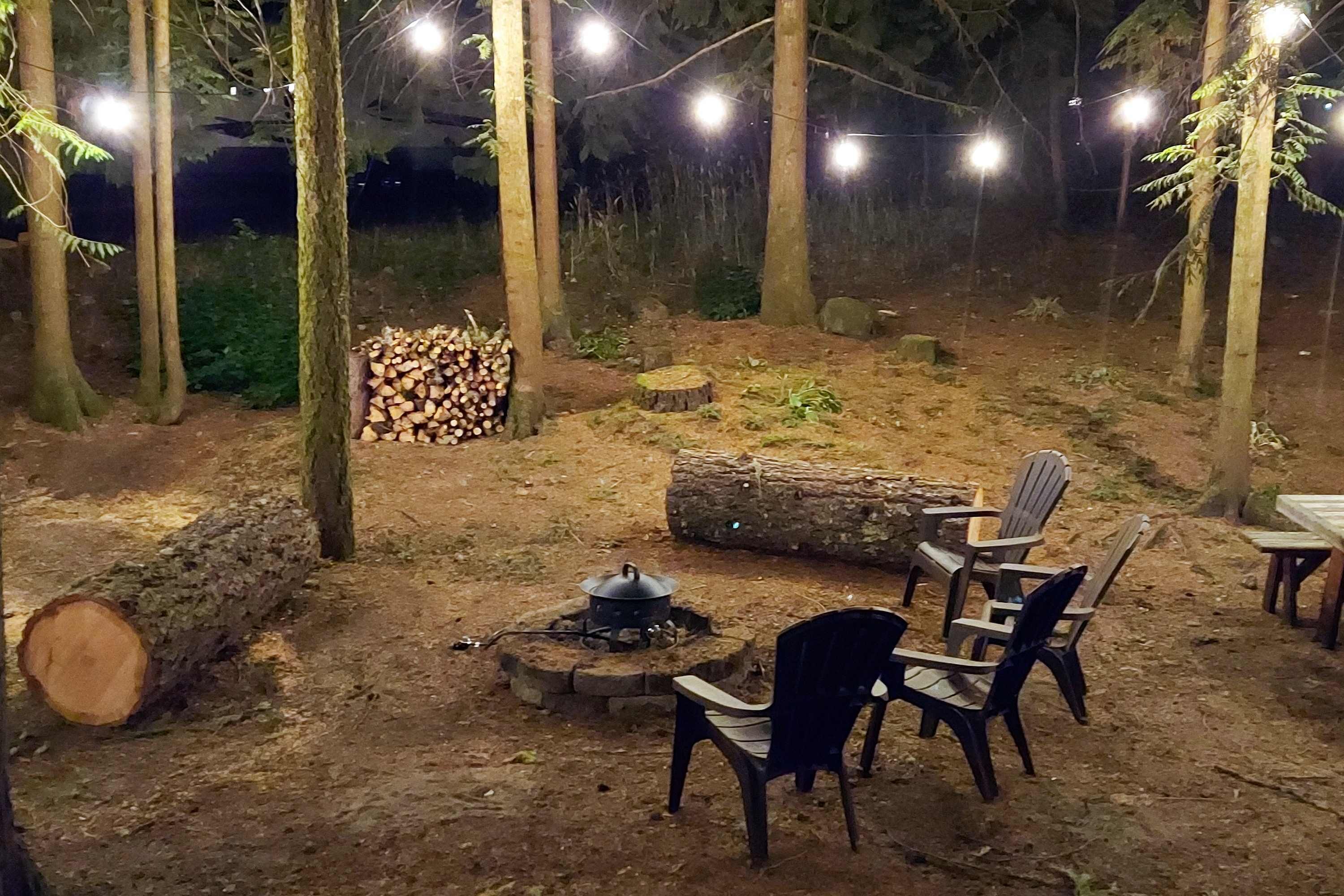 Een terras buiten met stoelen en een vuurkuil, omgeven door bomen en een stapel hout.