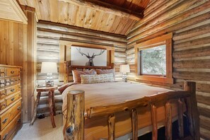 A log cabin bedroom with a bed, dresser, lamp, and a deer head mounted on the wall.