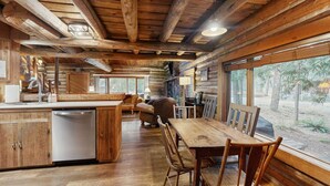 L’intérieur d’un chalet rustique est doté d’une table et de chaises en bois, d’un coin cuisine avec un lave-vaisselle et de grandes fenêtres offrant une vue sur l’extérieur.