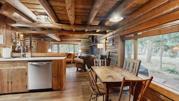 L’intérieur d’un chalet rustique est doté d’une table et de chaises en bois, d’un coin cuisine avec un lave-vaisselle et de grandes fenêtres offrant une vue sur l’extérieur.