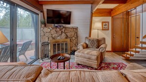 A cozy living room with a stone fireplace, a comfortable leather sofa, a television mounted on the wall, and a staircase leading to an upper level. - Cozy Family Cabin with Hot Tub, Bikes, Dog-Friendly, Free WIFI with SHARC passes (Sunriver)
