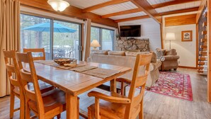 A wooden dining table with chairs, a stone fireplace, a television, and a sofa in a cabin-style room. - Cozy Family Cabin with Hot Tub, Bikes, Dog-Friendly, Free WIFI with SHARC passes (Sunriver)