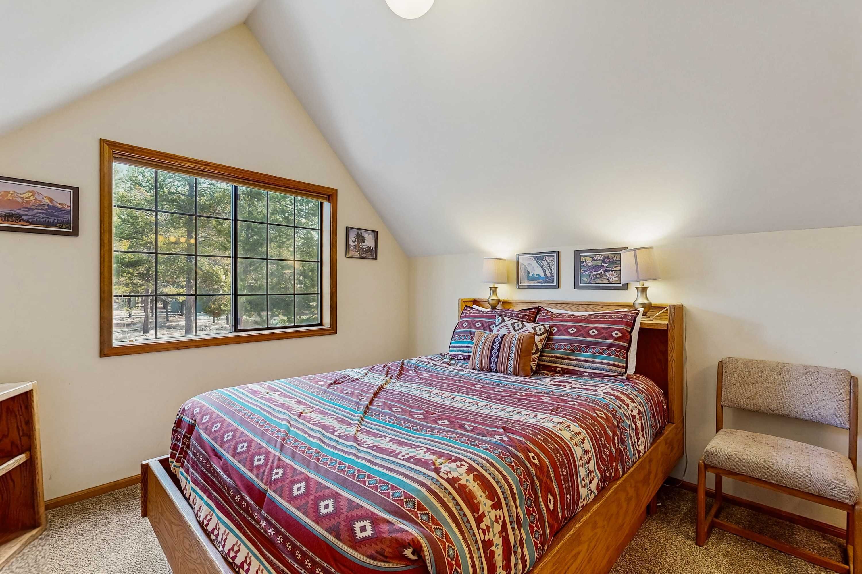 A bedroom with a large window, a bed with patterned bedding, a chair, and two wall-mounted lamps.