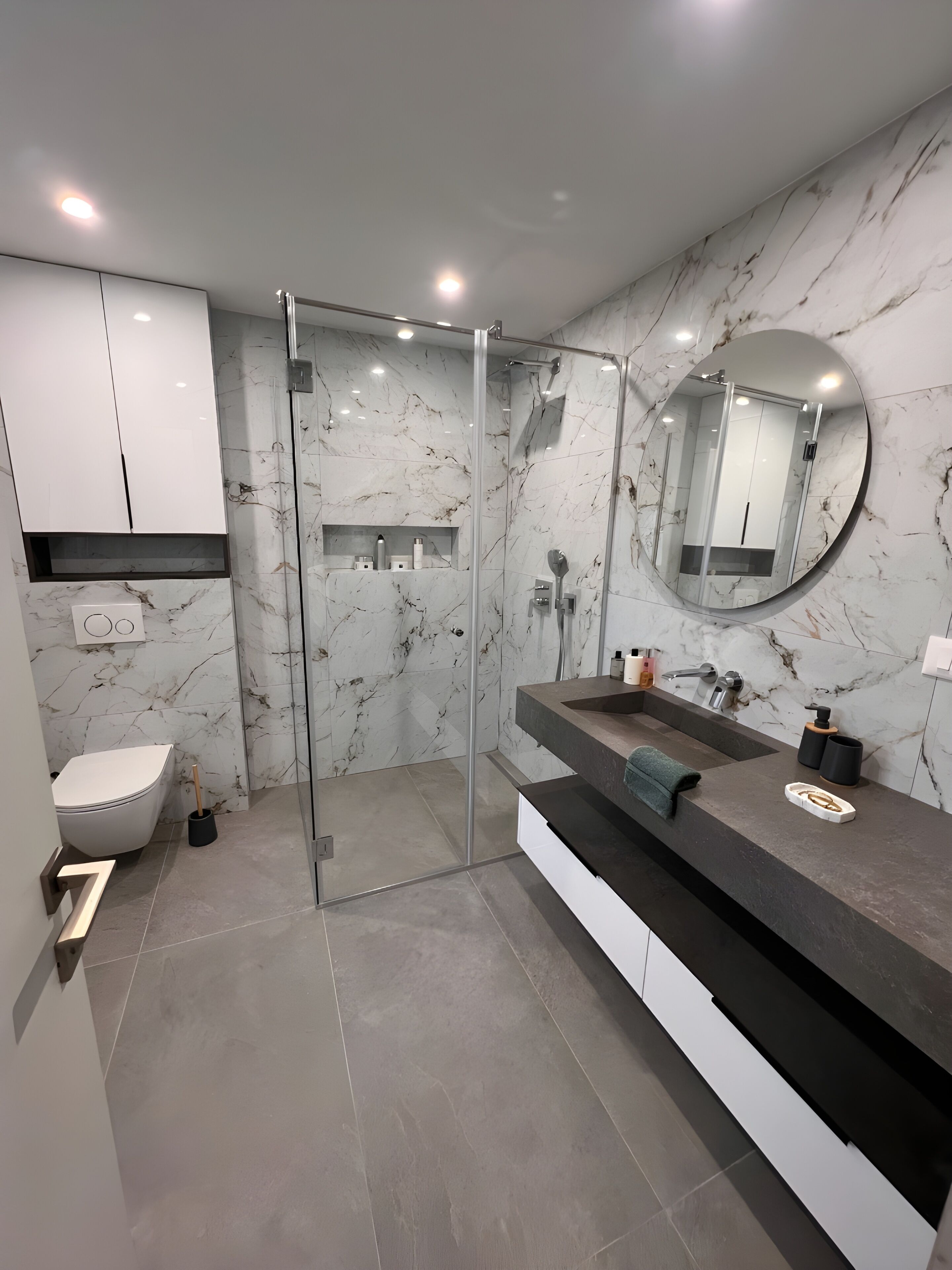 Un baño moderno con cabina de ducha de vidrio, pared con mosaicos de mármol, espejo redondo y tocador doble.