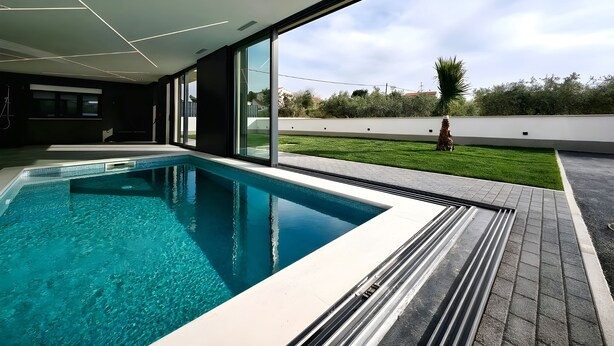 Ein moderner, überdachter Pool mit klarem, blauem Wasser, großen Fenstern und Blick auf eine gepflegte Rasenfläche.