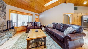 A cozy living room with a stone fireplace, leather sofas, a wooden coffee table, and large windows. - Dog-Friendly Sunriver Home with Fireplace, Deck & Central AC (Sunriver)