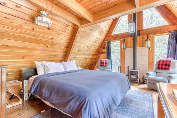 A wooden cabin bedroom with a bed, a chair, a table, a heater, and a window. - Intimate dog-friendly retreat with free WiFi, wood stove, & gas grill (Welches)