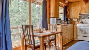 Interno di una baita in legno con tavolo da pranzo, sedie, angolo cottura e ampie finestre che si affacciano su una foresta.