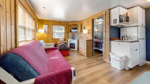 A cozy cabin with a bed, a small kitchenette, a fireplace, and a window with blinds. - Dog-Friendly Beach Condo with Free WiFi - Close to Beach (Cannon Beach)