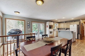 A kitchen with wooden cabinets, a dining table with chairs, a built-in island, and a view through the glass door. - Creekside cabin with water view, deck, jetted tub & game room - near Mt Hood (Rhododendron)