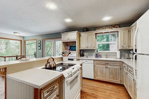 A kitchen with wooden cabinets, a stove, a dishwasher, and a window with a view of trees. - Creekside cabin with water view, deck, jetted tub & game room - near Mt Hood (Rhododendron)