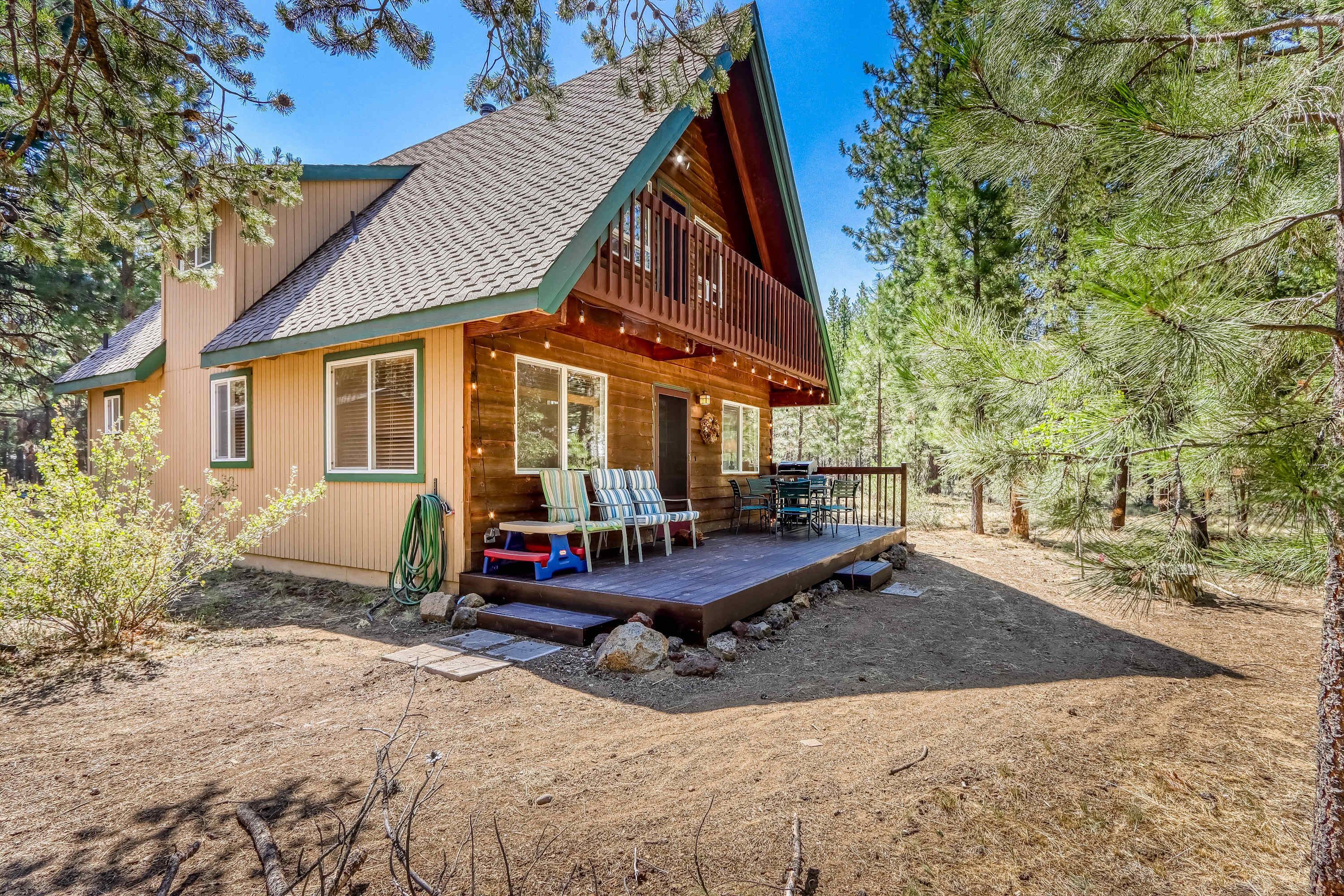 A log cabin with a deck, outdoor seating, and a dirt yard surrounded by trees.