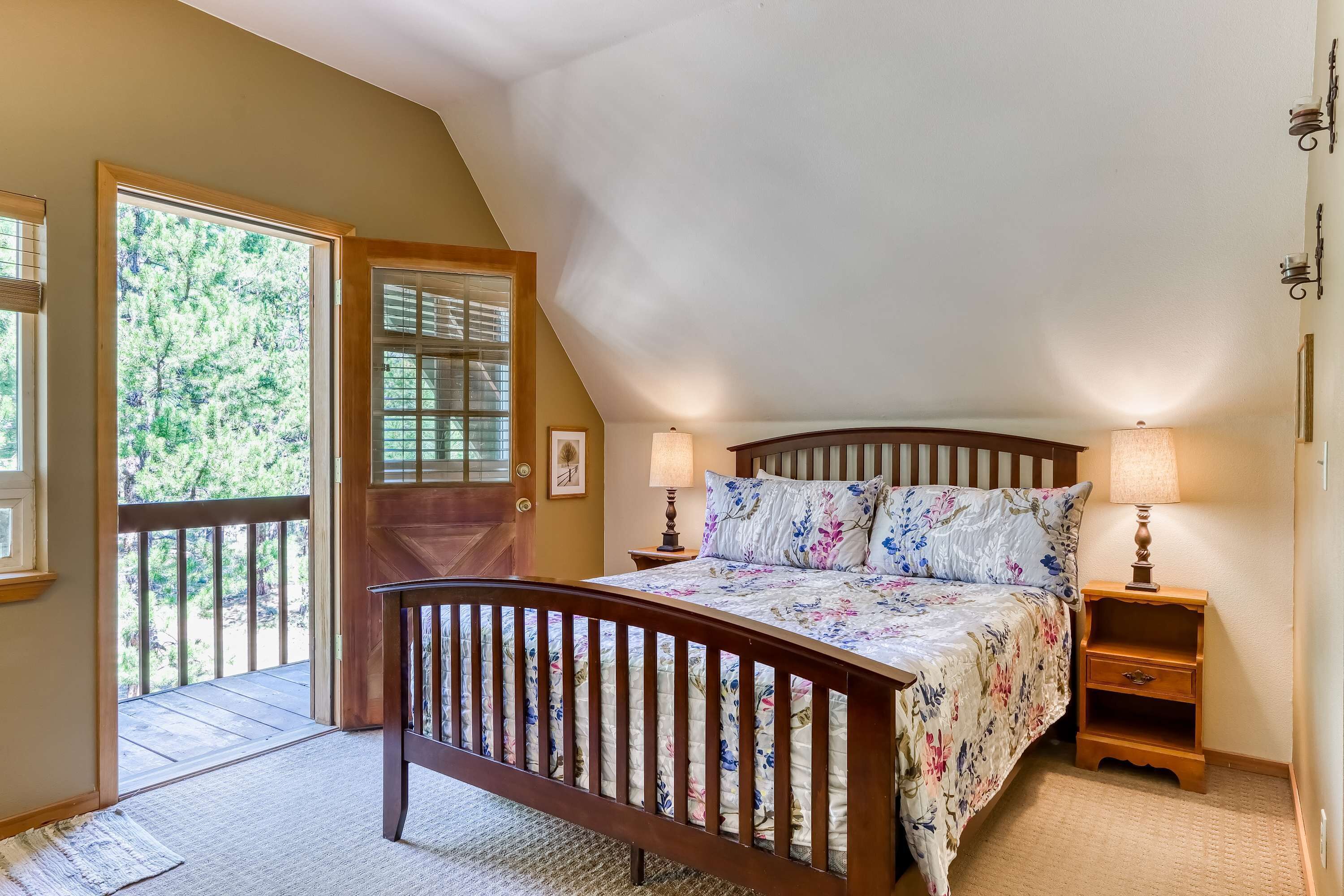 Una camera da letto con un letto in legno, due lampade sui comodini, un comodino in legno, una porta che dà su un balcone e vista sugli alberi.