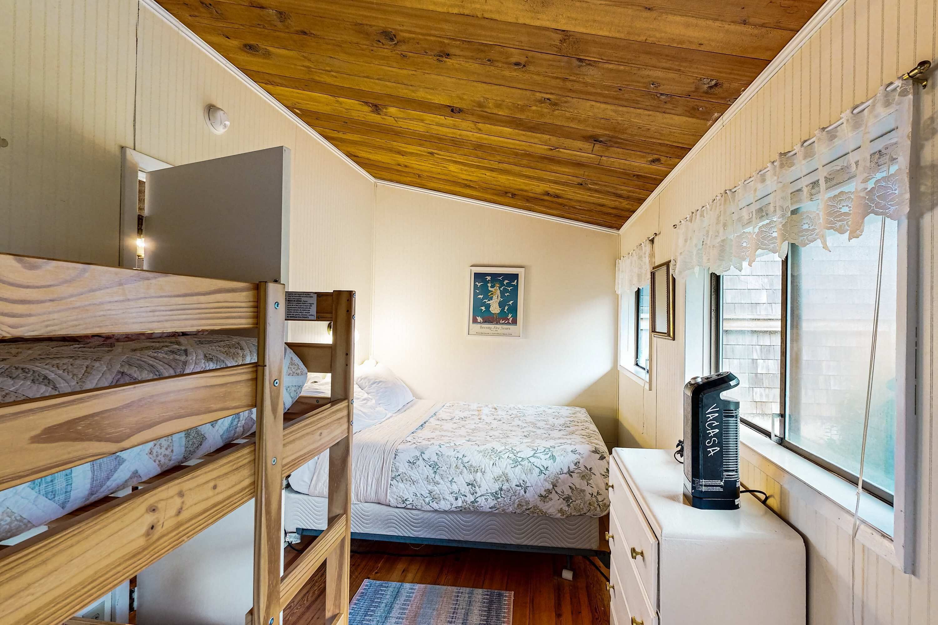 Una habitación con literas, una cama con ropa de cama floral, un escritorio pequeño con lámpara y una ventana con cortinas de encaje.