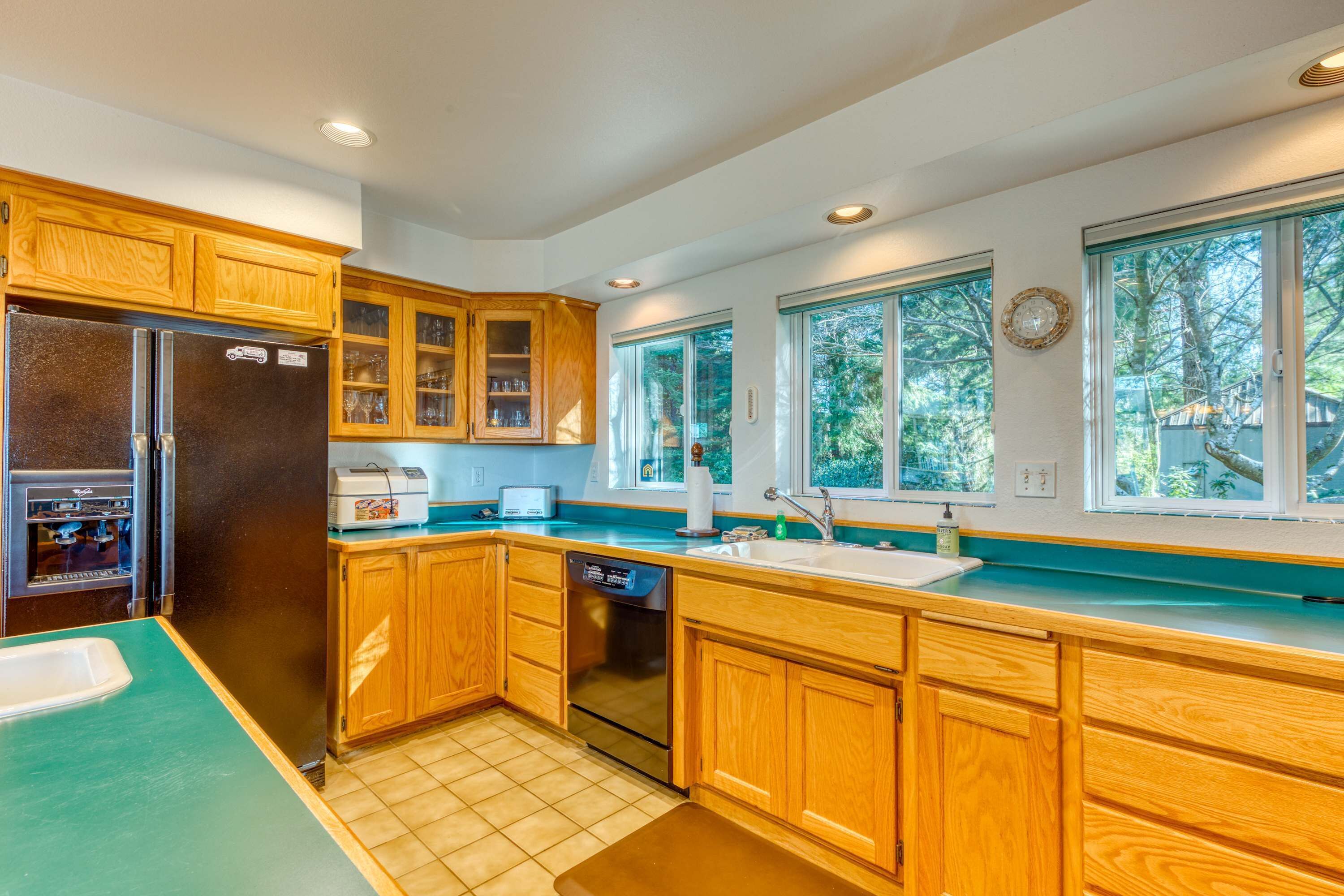 A kitchen with wooden cabinets, a refrigerator, a dishwasher, and a window with a view of trees.