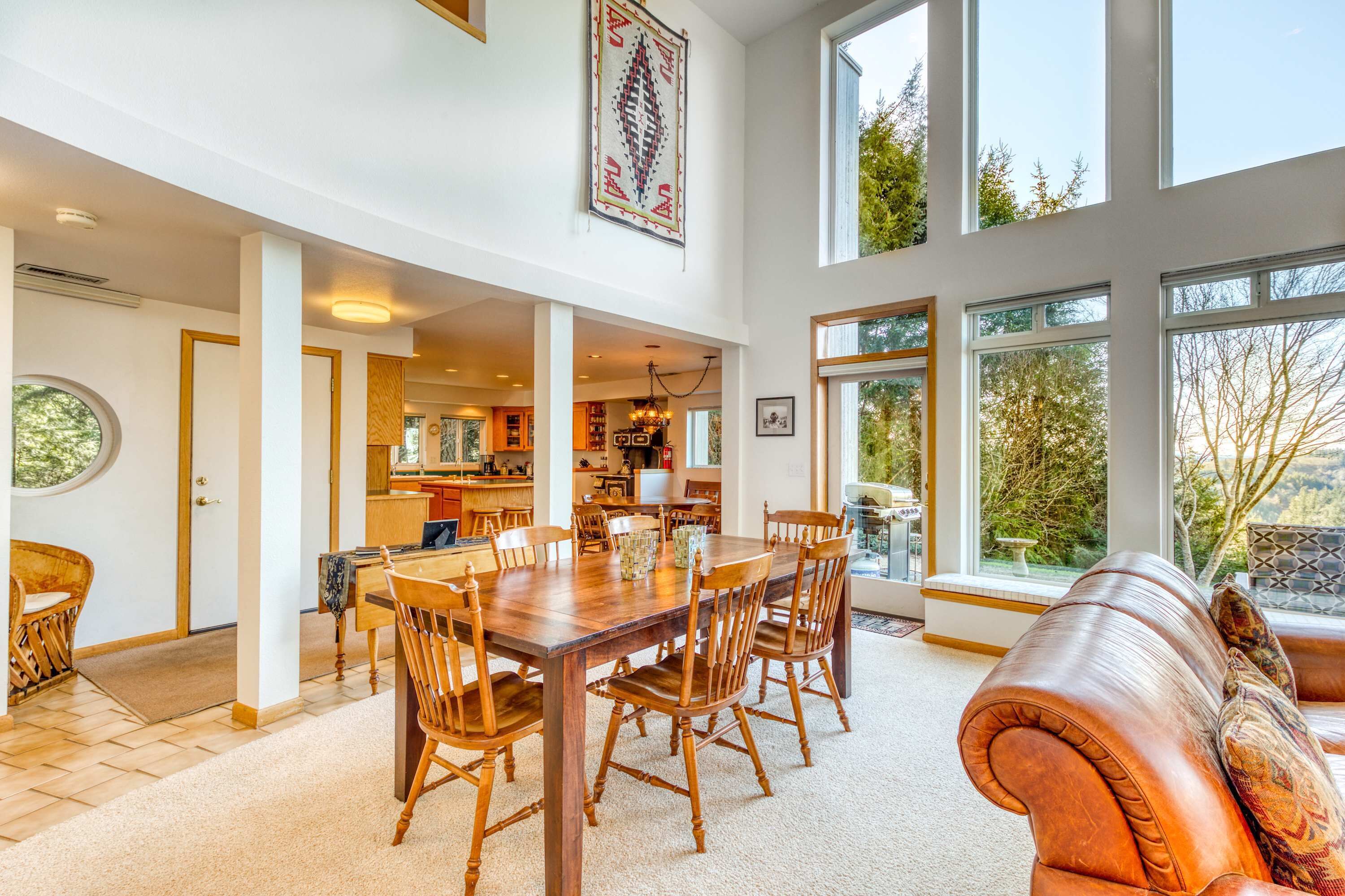 A spacious dining area with a large wooden table and chairs, a leather sofa, and large windows offering views of the outdoors.