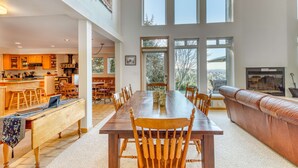 A spacious dining area with a long wooden table and chairs, a kitchen in the background, and large windows providing natural light. - Mid-century home with floor-to-ceiling windows & great views (Coos Bay)