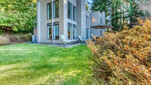 A modern two-story house with a well-maintained lawn, outdoor seating area, and a large umbrella. - Mid-century home with floor-to-ceiling windows & great views (Coos Bay)