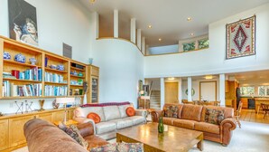 A spacious living area with multiple sofas, a coffee table, and a bookshelf filled with books and decorative items. - Mid-century home with floor-to-ceiling windows & great views (Coos Bay)