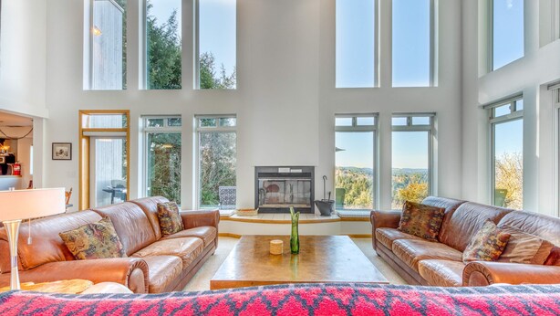 A spacious living room with leather sofas, a fireplace, and large windows offering views of the outdoors. - Mid-century home with floor-to-ceiling windows & great views (Coos Bay)