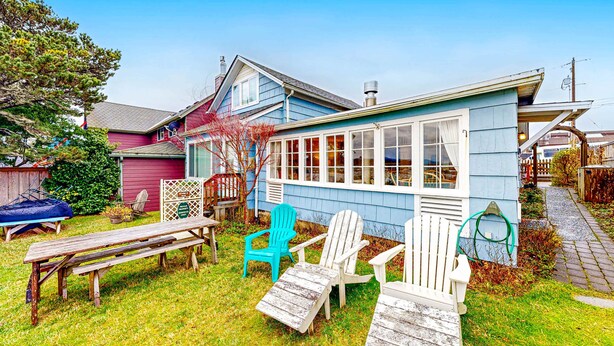 A house with a blue exterior, a covered porch, and a garden with a wooden bench and Adirondack chairs.