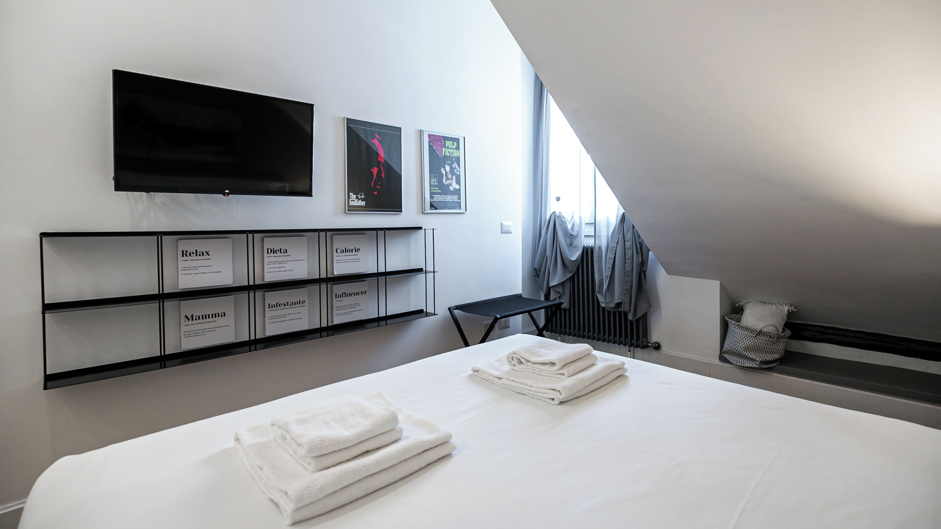 A modern hotel room with a large bed, a flat-screen TV mounted on the wall, and a shelf with informational cards.