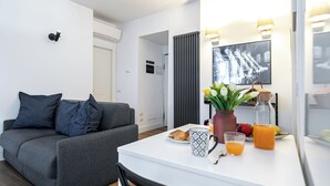 Een moderne woonkamer met een grijze bank, een eettafel met ontbijtartikelen, en een aan de muur bevestigde televisie.