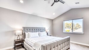 Une chambre à coucher avec une tête de lit capitonnée, une table de chevet avec une lampe, une grande fenêtre donnant sur un beau paysage et un ventilateur de plafond.