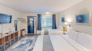 Hotellihuone, jossa on kaksi sänkyä, televisio, työpöytä, lamppu ja ikkuna, jossa on verhot.