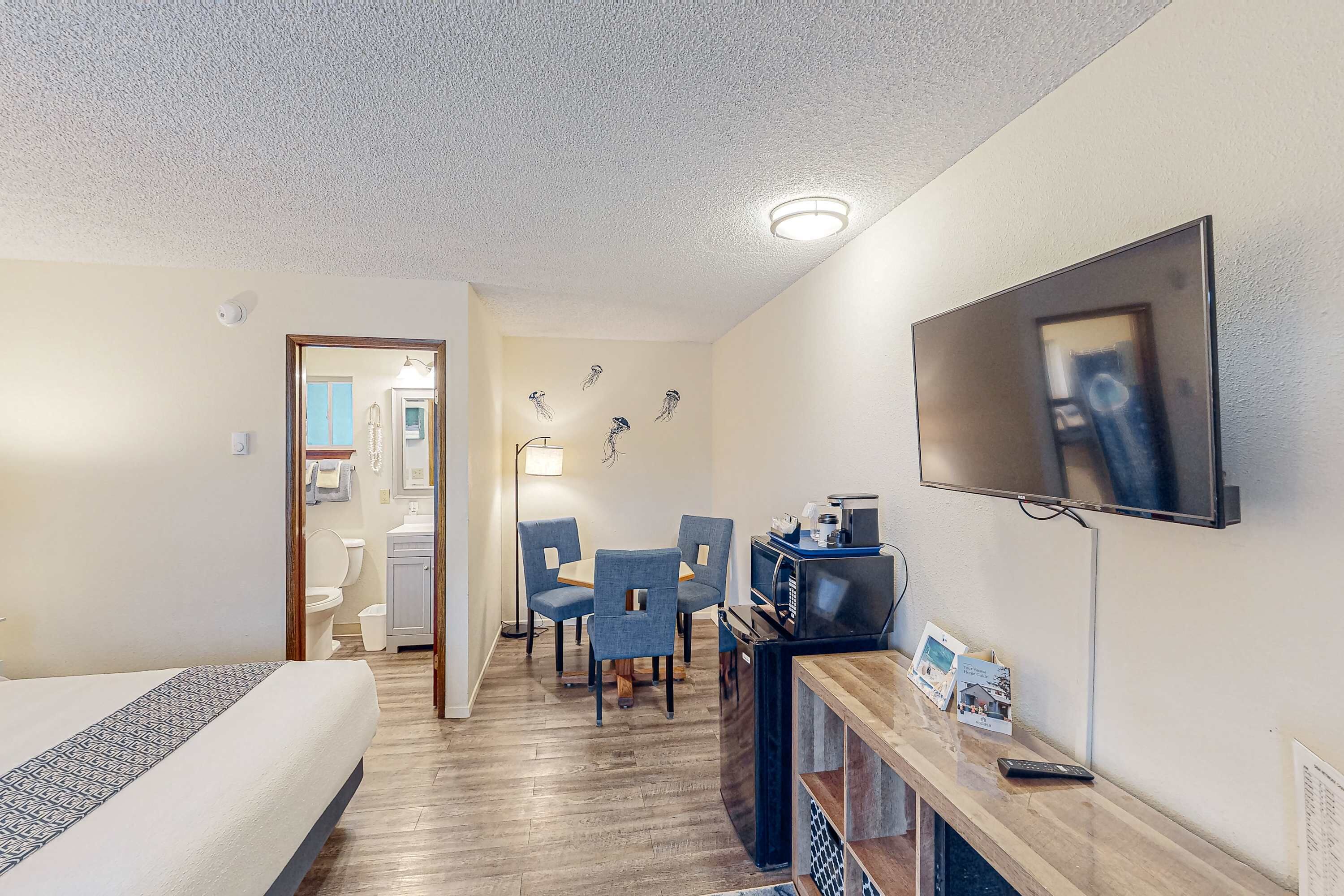 Habitación de hotel con cama, escritorio con televisor, zona de comedor con sillas y baño visible a través de una puerta abierta.