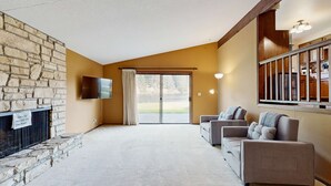 Una sala de estar con chimenea de piedra, un televisor de pantalla plana, un sofá y vistas al exterior a través de un ventanal.