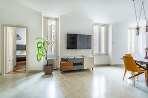 A modern living room with a flat-screen TV, a dining area with yellow chairs, a potted plant, and a large window. - iFlat | Stylish Crispi 20 Exclusive Apartment (Rome)