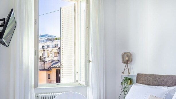 Hotellihuone, jossa on sänky, ikkuna, jossa on sälekaihtimet, yöpöytälamppu ja näkymä ulkona oleviin rakennuksiin.