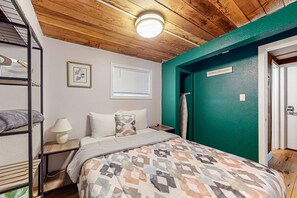 A room with bunk beds, a bed with a patterned bedspread, a bedside lamp, a window with blinds, and a door with a sign. - Dog-friendly rustic house near ocean and golf with washer/dryer & gas grill (Florence)