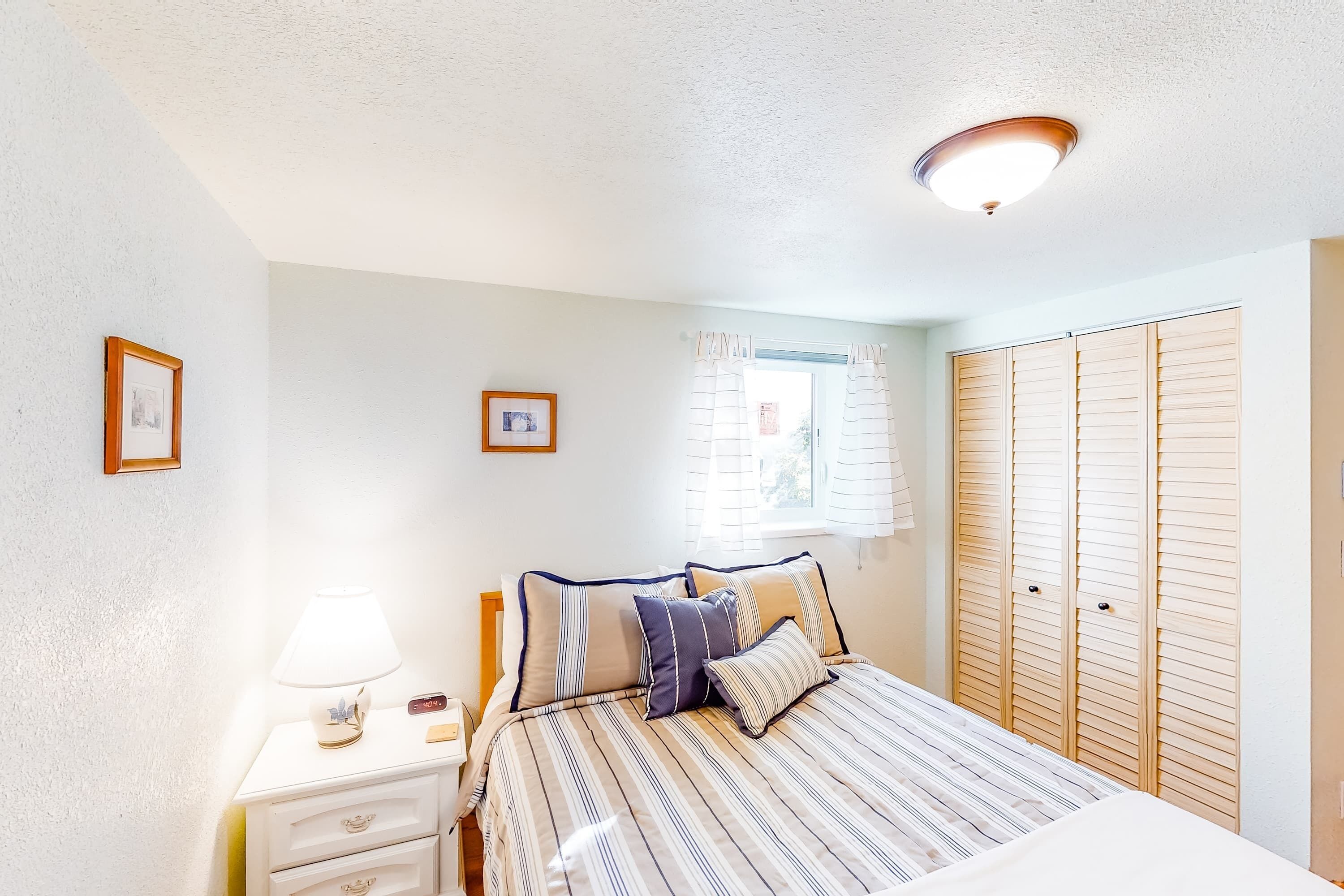 Een slaapkamer met een bed, nachtkastjes, een lamp, een raam met gordijnen en een kledingkast.