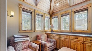 A cozy room with wooden walls, large windows, a small table, two patterned armchairs, and a lamp. - Spacious Family Home with Hot Tub, AC, Garage Gym & SHARC Amenities (Sunriver)