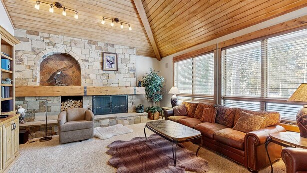 A cozy living room with a stone fireplace, wooden ceiling, and large windows. - Spacious Family Home with Hot Tub, AC, Garage Gym & SHARC Amenities (Sunriver)