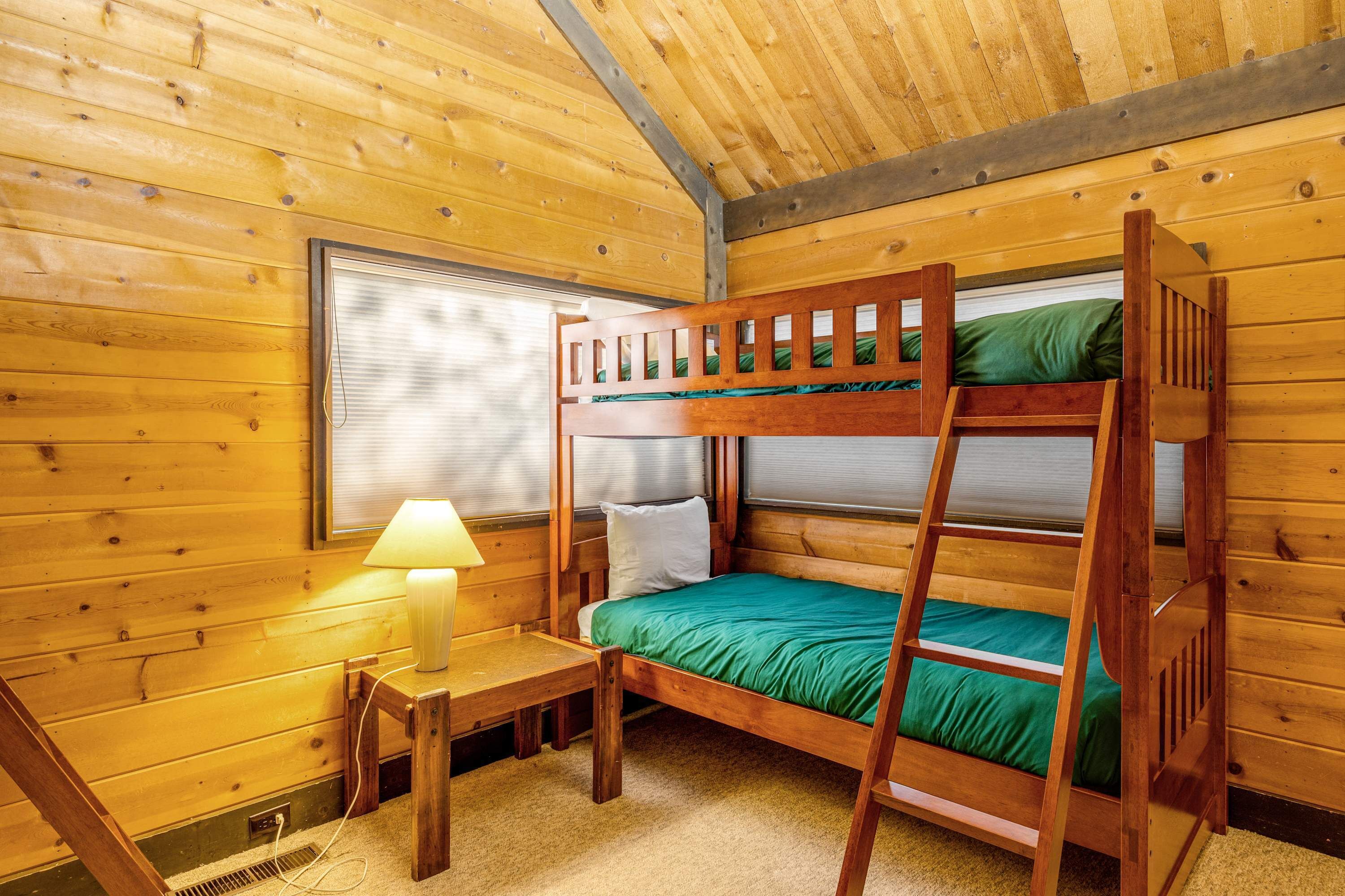 Quarto com beliches, paredes de madeira, janela, mesa com luminária e piso coberto por carpete.