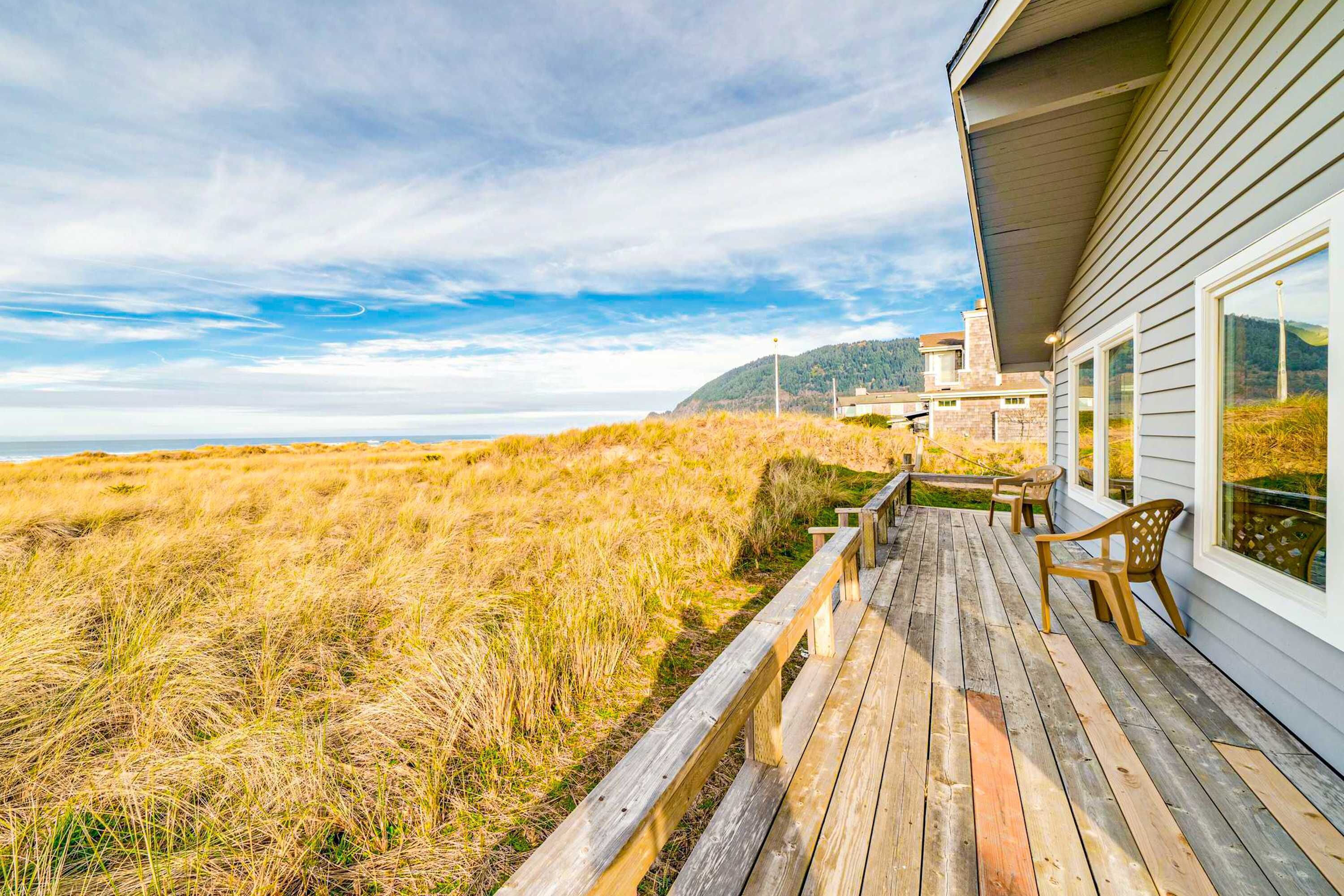 Eine Holzterrasse mit Sitzgelegenheiten im Freien, mit Blick auf einen Strand mit Dünen und eine ferne Bergsilhouette.