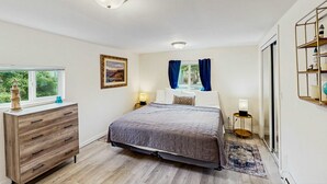 Un dormitorio con cama, cómoda, lámparas de noche, ventana con cortinas y vista a zonas verdes.