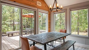 A dining area with a wooden table and benches, large windows, and a view of the forest. - Home with hot tubs, pools, tennis, pickleball, gym, fireplace, & game room (Black Butte Ranch)