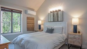 A bedroom with a large bed, two bedside tables with lamps, a wall-mounted air conditioner, and a window with a view of trees. - Home with hot tubs, pools, tennis, pickleball, gym, fireplace, & game room (Black Butte Ranch)