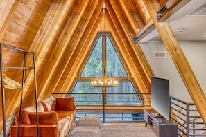 Une pièce aux hauts plafonds, avec un plafond en bois, une grande fenêtre, un canapé, un téléviseur et un lustre.