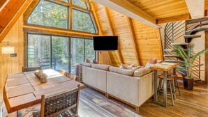 Ein gemütliches Blockhaus-Interieur mit großem Fenster, einer Couch, einem Esstisch und einer Wendeltreppe.