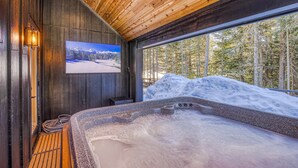 Ein Holzinnenraum mit einer Whirlwanne, einem Fernseher und Blick auf eine verschneite Landschaft.