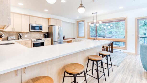 Une cuisine moderne dotée d’un grand îlot central, d’appareils électroménagers en acier inoxydable, de tabourets de bar en bois et de grandes fenêtres offrant une vue dégagée.